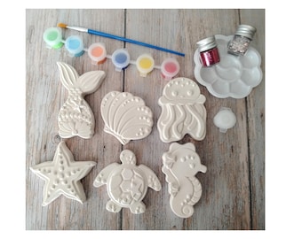 May include: A craft kit featuring various sea creature figurines, including a mermaid tail, starfish, and seahorse. The kit includes paint pots in various colors, a paintbrush, glitter, and a paint palette. The figurines are white and ready to be painted.
