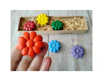 May include: A hand holds a bright orange flower-shaped crayon. Other flower crayons in red, yellow, green, blue, and purple are displayed on a wooden surface, some in a small cardboard box with shredded paper.