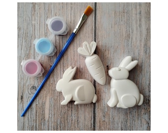 May include: An Easter-themed craft set featuring three small paint pots with purple, blue, and pink paint, a blue paintbrush, and three white ceramic shapes: two bunnies and a carrot. The items are arranged on a wooden surface.