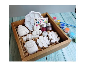 May include: A craft kit in a brown box, featuring bee-themed ceramic figurines, paint pots, glitter, a paintbrush, and a palette. The kit includes a beehive, bee, honey dipper, honeycomb, flower, and honey pot figurines.