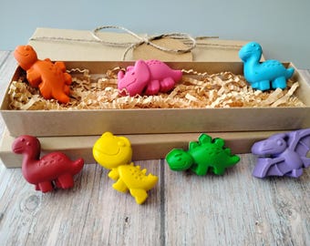 May include: A set of colorful dinosaur-shaped crayons in a kraft paper box with shredded paper filler. The crayons are in shades of orange, pink, blue, red, yellow, green, and purple. The box is tied with twine.