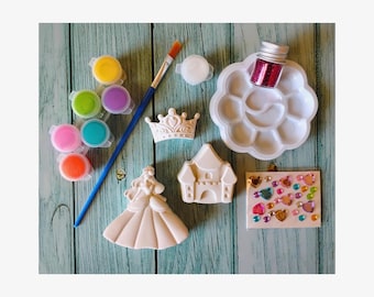 May include: A craft kit with paint pots, a paintbrush, a palette, and glitter. The kit includes ceramic figurines of a princess, a crown, and a castle, along with a small canvas decorated with colorful gems. The items are arranged on a light blue wooden surface.