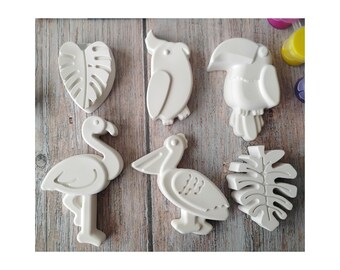 May include: A collection of white ceramic tropical-themed shapes, including a monstera leaf, parrot, toucan, flamingo, pelican, and another leaf. The shapes are arranged on a light brown wooden surface.