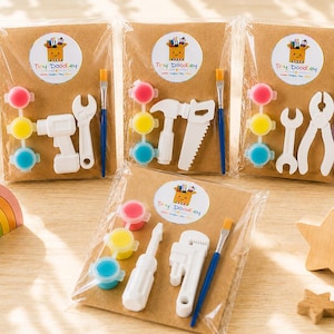 May include: Four craft kits in clear plastic packaging on brown cardstock, each featuring a Tiny Doodley logo, three small paint pots in pink, yellow, and blue, a blue-handled paintbrush, and two white plastic tool-shaped figures for painting.