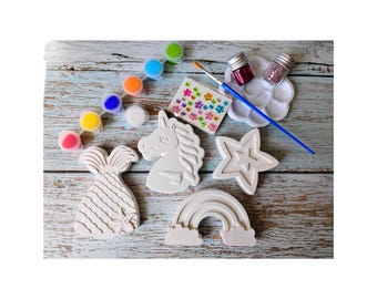 May include: A craft kit featuring white plaster figures of a mermaid tail, a unicorn head, a star, and a rainbow. Includes a strip of six paint pots, a blue paintbrush, a white palette with two glitter jars, and a sheet of colorful flower stickers.