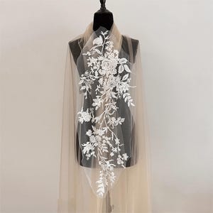 May include: A sheer beige tulle fabric panel featuring an elongated white floral lace appliqué. The intricate embroidery displays a vertical arrangement of roses, leaves, and small blossoms, centered on the translucent, lightweight mesh material.