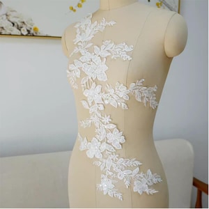May include: White floral lace applique with a delicate design of flowers and leaves. The applique is displayed on a mannequin, showcasing its shape and detail. Perfect for embellishing bridal wear or other garments.
