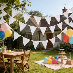 May include: A festive outdoor scene with white triangular bunting strung across a sunny backyard. Colorful balloons, a wooden table and chairs, and a picnic blanket suggest a celebration. A tiered dessert stand and a cottage in the background complete the cheerful setting.