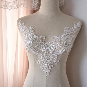 May include: A white lace applique featuring intricate floral and leaf embroidery with small bead accents. The decorative piece is shaped to fit the neckline and chest area, displaying a symmetrical pattern of swirls and petals on a beige mannequin.