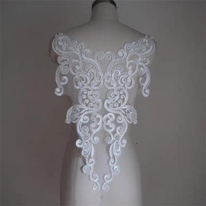 May include: A white lace applique featuring an intricate symmetrical scrollwork pattern with swirling floral motifs and delicate embroidery. The piece is displayed on a mannequin torso, showcasing its elongated shape and detailed stitching.
