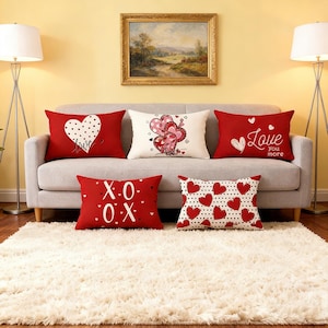 May include: A set of red and white decorative cushions on a grey sofa. The cushions display heart patterns, the words "Love you more", and the letters "X.O.X". The cushions are placed on a white shag rug.