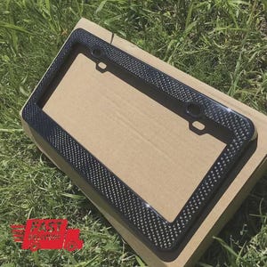 May include: A black license plate frame with a carbon fiber weave pattern. The frame is rectangular with rounded corners and mounting holes. The frame is sitting on a cardboard box, with green grass in the background.
