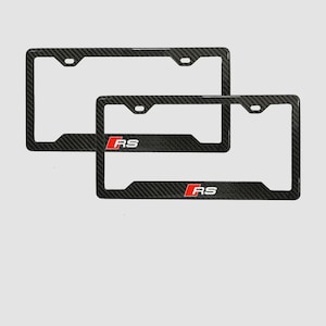 May include: Two black carbon fiber license plate frames. Each frame has a rectangular shape with rounded corners and the letters "RS" in red. The frames are designed to fit standard U.S. license plates.