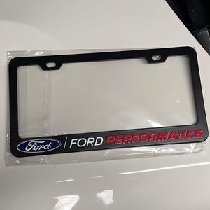 Puede incluir: Marco negro para matrícula con el logotipo de Ford y las palabras "FORD PERFORMANCE" en blanco y rojo. El marco está en un embalaje de plástico transparente.