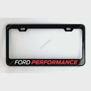 Könnte beinhalten: Schwarzer Kennzeichenrahmen mit den Worten "FORD PERFORMANCE" in weißer und roter Schrift. Der Rahmen hat eine rechteckige Form mit abgerundeten Ecken und Befestigungslöchern oben.