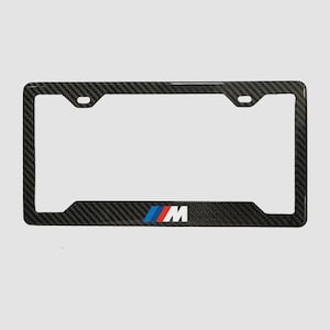 Könnte beinhalten: Schwarzer Kennzeichenrahmen aus Kohlefaser mit dem BMW M-Logo in Rot, Blau und Weiß. Der Rahmen hat eine rechteckige Form mit Befestigungslöchern und eine glänzende Oberfläche. Für Standardkennzeichen.