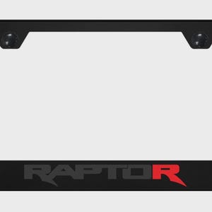 May include: Black license plate frame with the word "RAPTOR" in gray and red lettering. The frame has a rectangular shape with rounded corners and two small black mounting points at the top.
