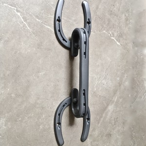 May include: A gray metal horseshoe-shaped wall hook. The hook is made of two horseshoe shapes connected by a vertical bar. The hook is mounted on a light gray marble-like surface.
