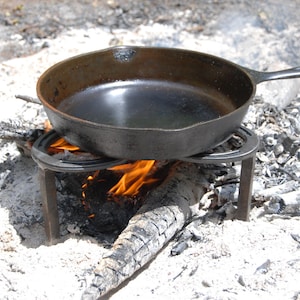 May include: A black cast iron skillet sits on a metal tripod over a small campfire. The skillet is empty and the flames are visible beneath it.