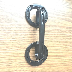 May include: Black metal door pull with a horseshoe design. The pull features two circular elements with horseshoe cutouts, connected by a vertical handle. The hardware is mounted on a light brown wooden surface.
