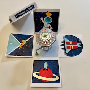 May include: A pop-up space-themed card with various space elements. The card features an astronaut, a rocket, a planet with a ring, and a spaceship. The card is white with blue, red, yellow, and silver accents. The box reads "Rockets & Stars".