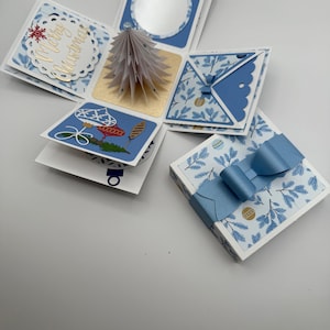 May include: A handmade Christmas explosion box with blue and white patterned paper. The box features a pop-up paper Christmas tree, decorative ornaments, and the words "Merry Christmas" in gold script. A blue bow adorns the closed box.