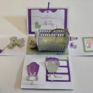 May include: A handmade baby-themed greeting card with a pop-up crib, a teddy bear, and decorative elements. The card features the words "welcome baby" and includes cut-out images of a duck, a baby, and clothing. The color scheme is primarily purple, green, and white.