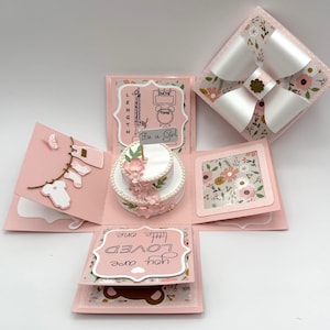 May include: A pink, multi-layered explosion box with a baby shower theme. It features a small cake, floral accents, and the phrase "It's a Girl." The box includes a large bow and decorative elements.