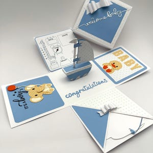 May include: A blue and white baby-themed explosion box. The box features a sailboat, a teddy bear, and a duck. The box has the words "welcome baby", "congratulations", and "It's a boy" on it. The box is decorated with white bows.