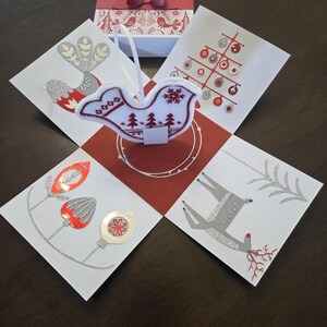 May include: A festive Christmas-themed gift box with a red and white bird ornament. The box is surrounded by cards with holiday illustrations of birds, trees, and deer. The box is tied with a red ribbon.
