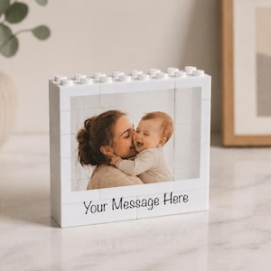 Puede incluir: Marco de fotos blanco en forma de bloque con una foto de una madre besando a un bebé. El marco tiene un diseño tipo Lego y el texto "Your Message Here" en la parte inferior. Un jarrón con vegetación está en el fondo.