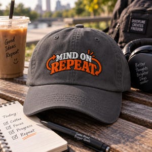May include: A charcoal grey baseball cap featuring the embroidered text "MIND ON REPEAT" in white and orange. The cap has a curved brim and a structured crown with ventilation eyelets, showcasing a clean design suitable for casual wear.
