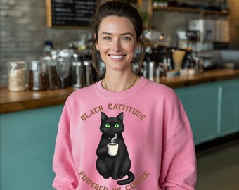 May include: A pink sweatshirt featuring the text "BLACK CATTITUDE POWERED BY COFFEE" and a black cat graphic with green eyes, holding a coffee cup. The sweatshirt is worn by a person.