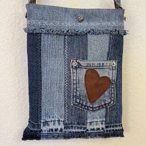 May include: A small denim crossbody bag with a brown leather heart patch on the front pocket. The bag has a frayed edge and a silver chain strap.