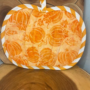 May include: A pumpkin-shaped quilted pot holder featuring an orange fabric with a repeating pattern of pumpkins and leaves. The edges are finished with a white and orange diagonal striped binding, and a matching fabric loop is attached at the top.
