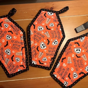 May include: Three coffin-shaped pot holders with a black polka dot trim. The orange fabric is patterned with white skulls, coffee cups, bats, and the text "Nightmare Before Coffee". Each has a black loop for hanging.