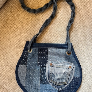 May include: A small, round, blue denim bag with a braided denim strap. The bag is made of patchwork denim and has a heart-shaped pocket on the front.