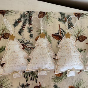 May include: Five white Christmas tree ornaments with gold glitter and star accents. Each tree has a ribbon for hanging and a small bow at the base. The ornaments are on a festive patterned background.