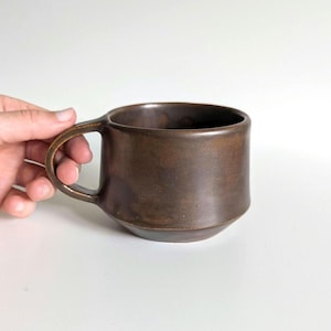 Puede incluir: Una taza de cerámica marrón con un asa redondeada. La taza tiene un cuerpo cilíndrico con una base ligeramente ensanchada. La taza es sostenida por una mano sobre un fondo blanco.