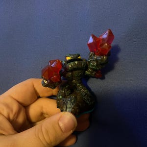 May include: A black and green action figure with two large red crystal-like structures. The figure has a detailed, textured design and a small yellow-eyed face. The figure is held in a hand against a blue background.