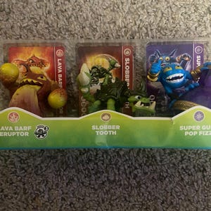May include: A plastic package containing three action figures: Lava Barf Eruptor, Slobber Tooth, and Super Gulp Pop Fizz. Each figure is displayed with a character card. The package has a green base with character names.
