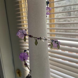 Puede incluir: Una delicada guirnalda floral hecha a mano con cuentas de flores moradas translúcidas, pequeñas cuentas rojas y dijes de hojas verdes. La guirnalda está ensartada en un alambre plateado e incluye una cuenta de cristal transparente. Para fines decorativos.