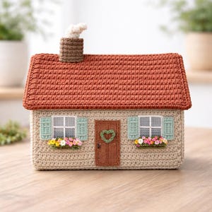 May include: A crocheted house with a terracotta roof, beige walls, and a brown door. The house features green shutters, white-framed windows, and window boxes filled with colourful flowers. A chimney with smoke detail completes the design.