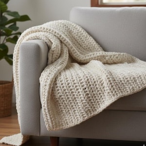 May include: A cream-colored, chunky knit blanket draped over the arm of a gray sofa. The blanket has a thick, textured weave, creating a cozy and inviting look. The neutral color palette complements the modern aesthetic of the room.