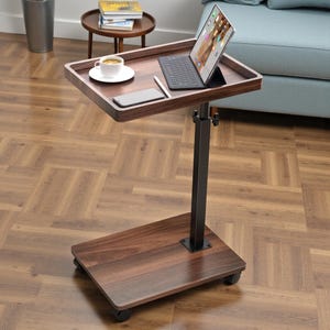 May include: A brown wooden laptop table with a black metal adjustable stand and a rolling base. The table surface holds a tablet with a keyboard, a smartphone, a pen, and a cup of coffee. The table is on a hardwood floor.
