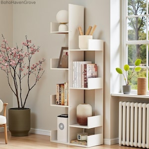 May include: A white, modern, tree-shaped bookshelf with multiple shelves. The shelves hold books, decorative vases, a framed picture, and a small lamp. A potted plant with pink flowers is to the left.