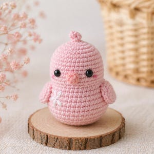 Könnte beinhalten: Ein handgehäkeltes rosa Küken mit schwarzen Sicherheitsaugen und einem weißen Blumendetail. Das Küken sitzt auf einer kleinen Holzscheibe. Ein Weidenkorb und rosa Blüten befinden sich im Hintergrund.