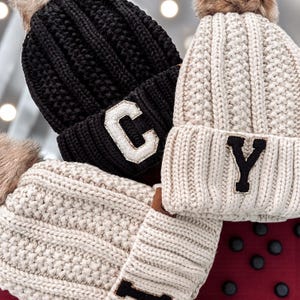 Puede incluir: Primer plano de dos gorros de punto, uno negro y otro color crema, cada uno con un parche de letra. El gorro negro tiene un parche 'C' blanco, mientras que el gorro crema tiene un parche 'Y' negro. Ambos gorros tienen un pompón de piel sintética.