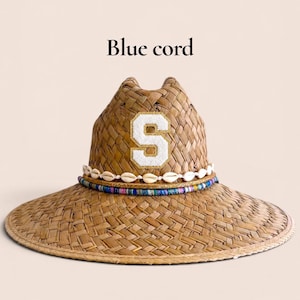 Puede incluir: Un sombrero de paja trenzada con un parche de la letra S blanca con un borde de purpurina dorada en la parte delantera. El sombrero está decorado con una banda de conchas cauri blancas, un cordón dorado fino y un cordón azul multicolor. Texto Blue cord.