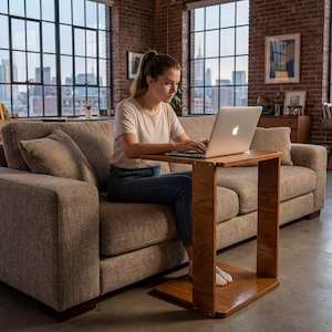 Può includere: Un tavolino in legno per laptop con piano e base rettangolari, collegati da due supporti verticali. Il tavolino è posizionato davanti a un divano beige. Una persona sta usando un laptop sul tavolino. La stanza ha grandi finestre e un muro di mattoni.